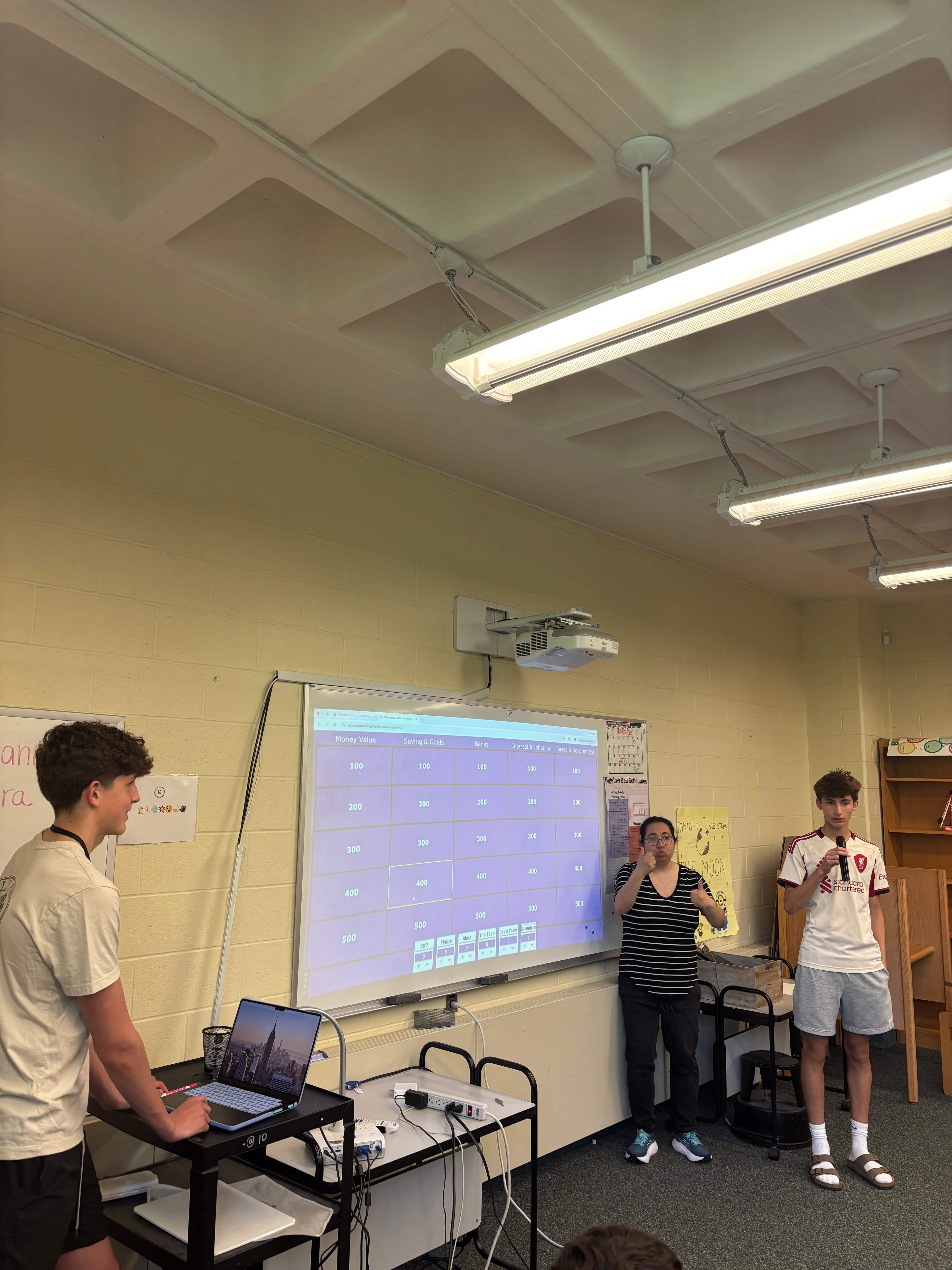 FLY presenters running a money-themed Jeopardy review game with eighth graders, the board projected on screen.