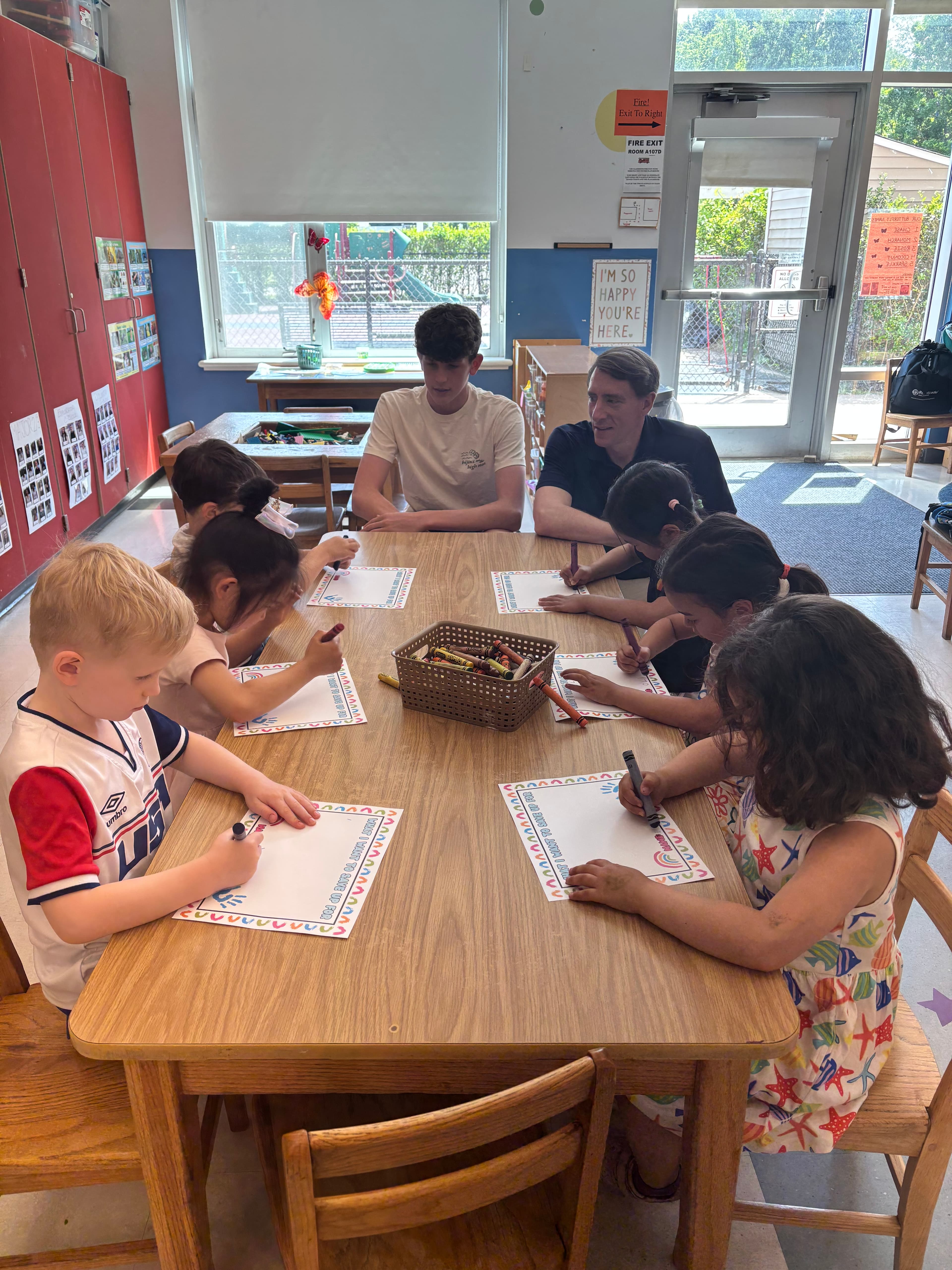 Two FLY volunteers leading a money activity with preschoolers coloring worksheets at a classroom table.