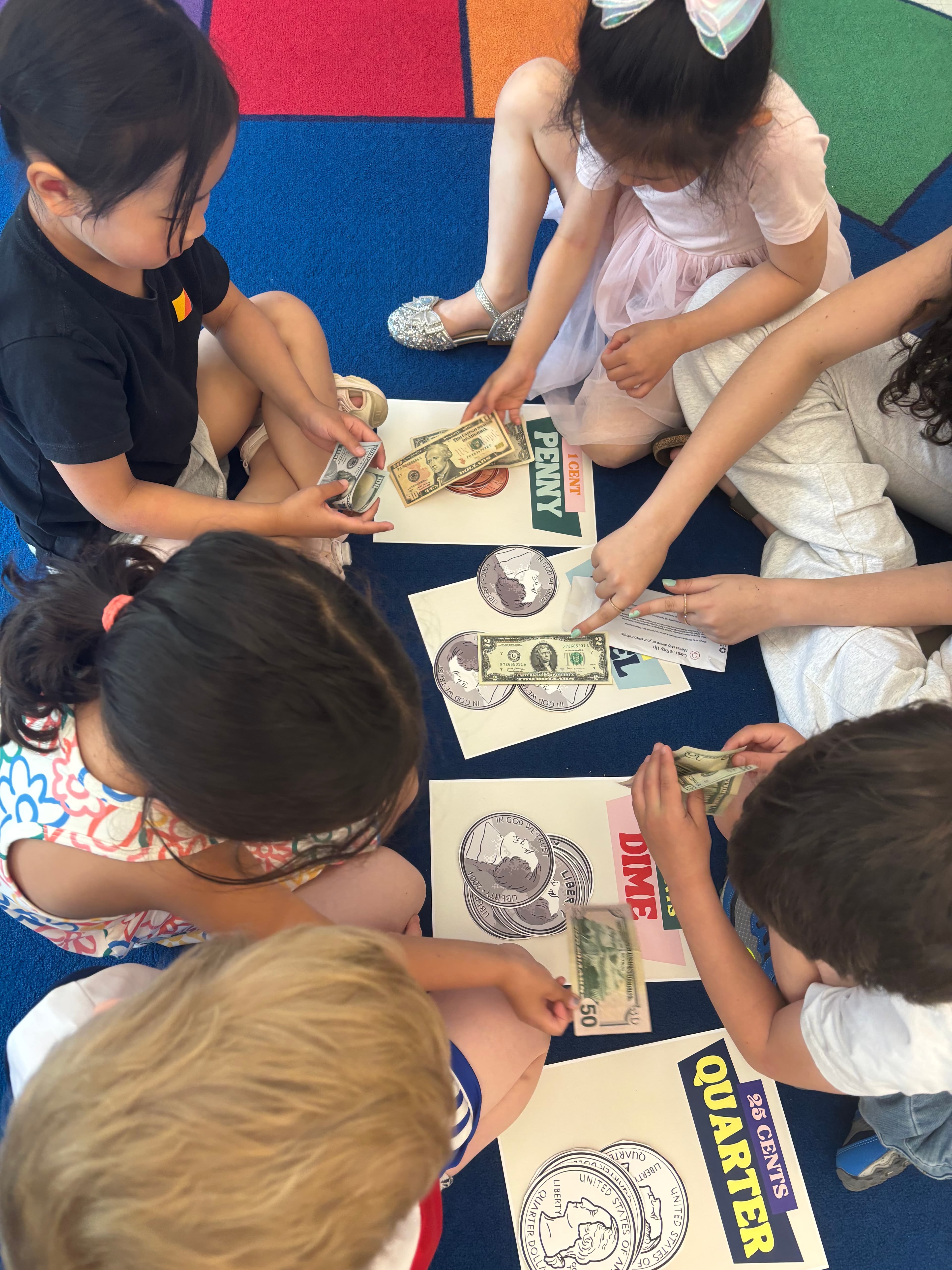 Preschoolers sorting coins and bills onto labeled penny, dime, and quarter cards on the floor.